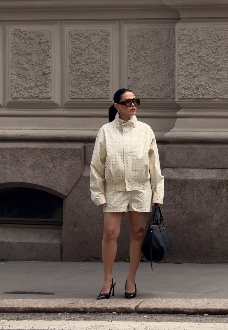 Femme portant une veste crème à pois et un short, talons hauts noirs, lunettes de soleil, tenant un sac à main noir, debout sur un trottoir urbain.