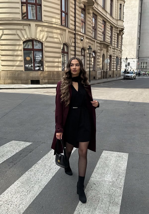 Žena s dlhými vlnitými vlasmi kráča po priechode pre chodcov, oblečená v čiernych šatách, bordovom kabáte, čiernych topánkach a nesie čiernu kabelku v mestskom prostredí.