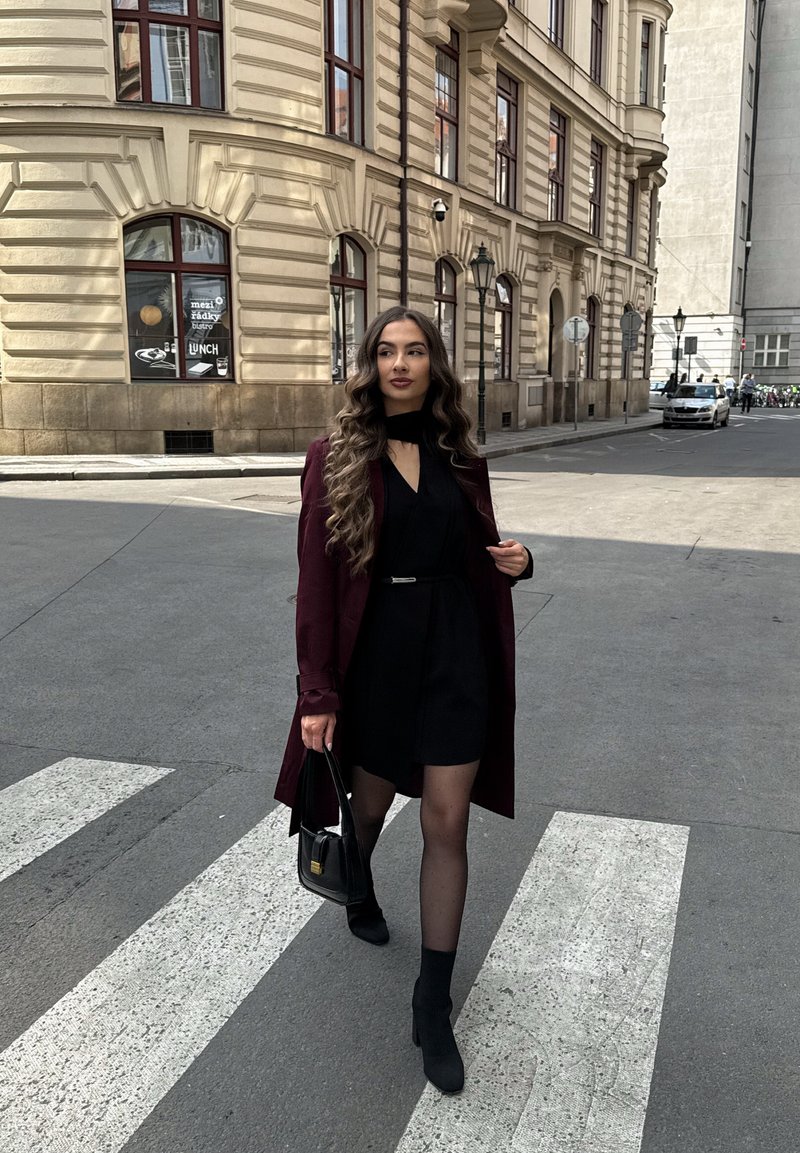 Femme aux longs cheveux ondulés marche sur un passage piéton portant une robe noire, un manteau bordeaux, des bottes noires et portant un sac à main noir dans un cadre urbain.