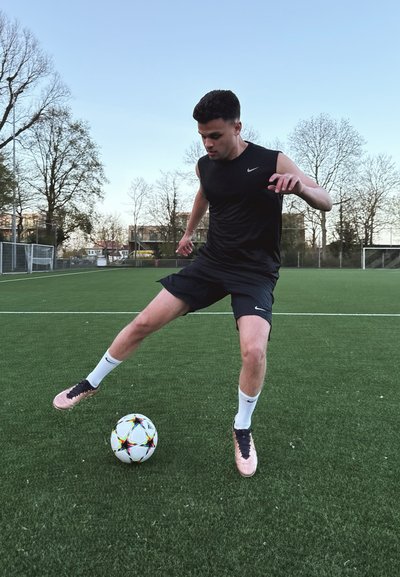 Atleta en camiseta sin mangas negra y pantalones cortos realiza un movimiento técnico sobre un balón de fútbol. Fondo de campo de hierba con árboles. Zapatos de fútbol rosas y negros.