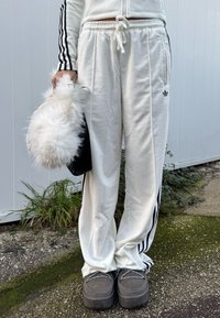 Άτομο που φοράει λευκό παντελόνι φόρμας Adidas και λευκό μπουφάν, κρατώντας μια αφράτη λευκή τσάντα, στέκεται έξω σε σκυρόδεμα.