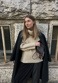 Beige bordázott garbós pulóver, fekete kabát, fekete csíkos nadrág, egy kicsi barna bőr táska tartása, textur�ált kőfal háttér.