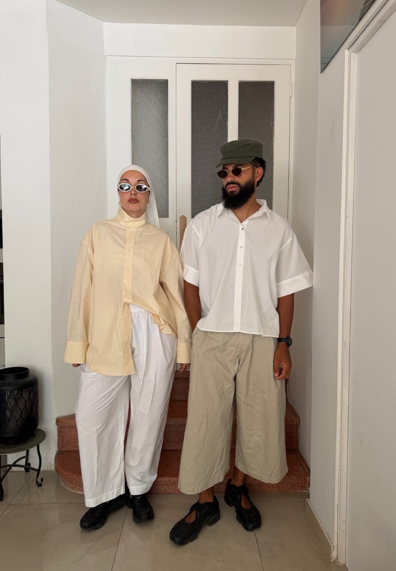 Deux personnes se tiennent à l'intérieur près des escaliers, vêtues de vêtements amples et clairs, de lunettes de soleil et de chaussures noires ; l'une porte un foulard, l'autre une casquette verte.