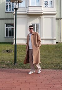 Bruine wollen overjas, witte trui, beige broek en zwart-witte sneakers. Staande op bakstenen trottoir nabij groen gras en een gebouw.