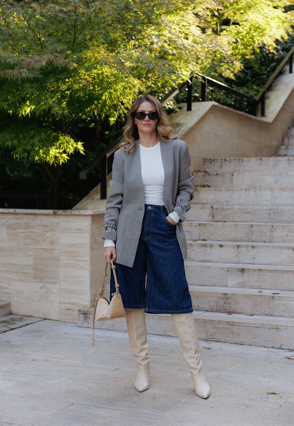 Blazer oversize gris, top blanc, culottes en denim foncé, bottes beiges jusqu'aux genoux et sac à main assorti. Des lunettes de soleil noires complètent le look.