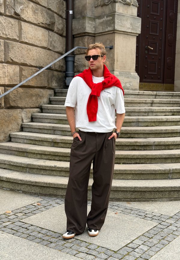 Un hombre con gafas de sol, una camiseta blanca y pantalones anchos está de pie en unos escalones de piedra, con un jersey rojo sobre los hombros.