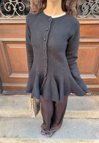 Cardigan péplum en laine noire avec détails à boutons, manches slim, texture douce, associé à un pantalon marron et un sac matelassé.