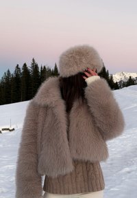 Manteau en fausse fourrure beige clair, texturé avec un large col. Associé à un pull en tricot beige et un chapeau en fourrure assorti. Fond enneigé.