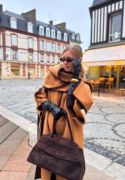 Capa marrón de lana con diseño en capas, guantes de cuero negros y un bolso oscuro de ante. Unas pequeñas gafas de sol oscuras completan el conjunto.