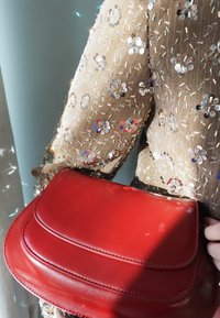 Rød læderhåndtaske med en glat finish, der har lagdelte frontklapper. Båret med en beige beklædning med pailletter og udsmykket med blomsterprint.