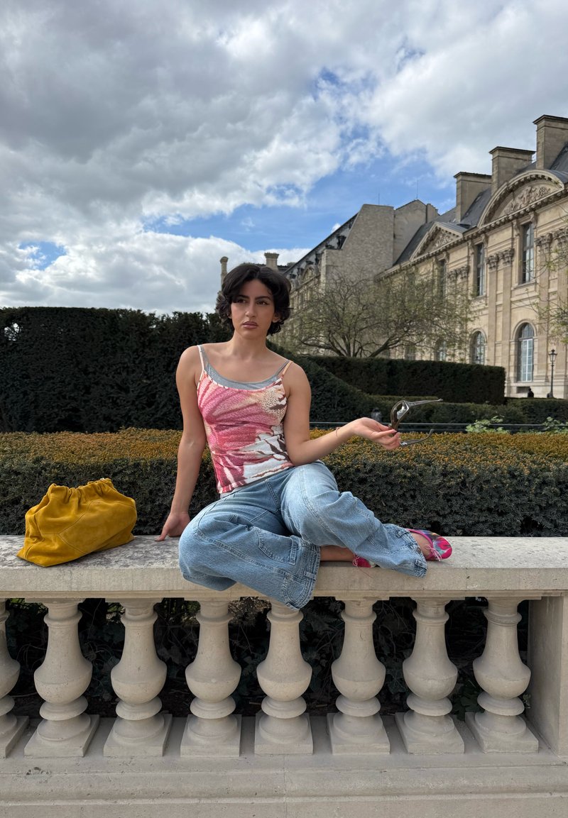 Jeune personne aux cheveux courts et foncés assise en tailleur sur une balustrade en pierre près d'une haie et d'un bâtiment classique, tenant des lunettes de soleil et se reposant à côté d'un sac jaune.