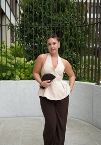 Beige halterneck-top dengan detail kancing, pinggang peplum, dan celana lebar cokelat tua, memegang clucth bertekstur gelap. Latar belakang dedaunan hijau.