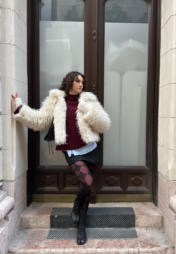 Chaqueta de piel sintética cremosa y mullida, suéter burdeos, medias negras y rojas con estampado, y zapatos negros, de pie en las escaleras junto a una puerta marrón.