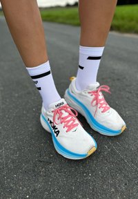 White running shoes with pink laces and a blue midsole, featuring a textured fabric upper and a black logo. Paired with white socks with black accents.