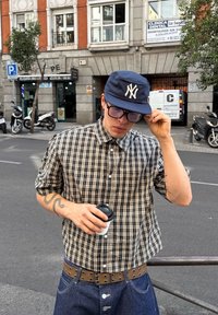 Čierno-biele kárované tričko s krátkym rukávom, tmavomodrá čiapka s logom, džínsy, hnedý opasok s kovovými hrotmi, drží kávový pohár.