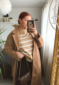 Brun lång kappa över en beige och vitrandig tröja, ihop med svarta byxor och en liten rutigt väska. Inomhusmiljö, naturligt ljus.