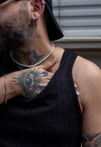 Black ribbed tank top, silver hoop and cross earrings, layered pearl necklace, intricate hand tattoo featuring an eye, and patterned skin texture.