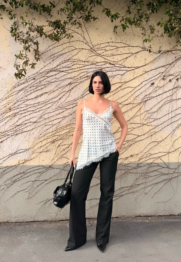 Femme aux cheveux courts et foncés portant un haut blanc à pois avec bordure en dentelle et un pantalon noir, tenant un sac à main noir, debout contre un mur beige couvert de vigne.