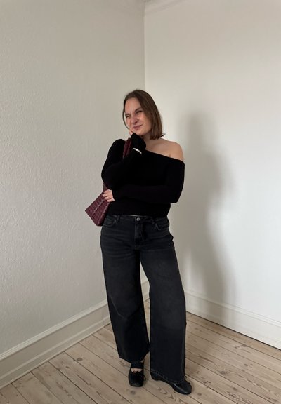 Mujer con top negro sin hombros, vaqueros negros anchos y zapatos negros posa en interiores, sosteniendo un bolso texturizado de color rojo oscuro sobre su hombro.