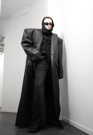 Persona con abrigo negro de cuero oversized, pañuelo negro en la cabeza, gafas de sol, pantalones negros de pierna ancha, de pie en una esquina blanca en interiores.