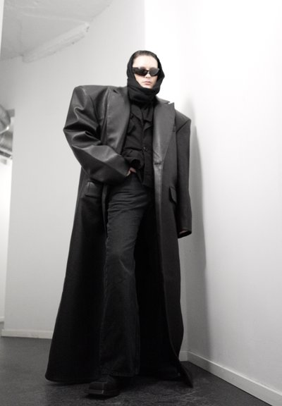 Persona con abrigo negro de cuero oversized, pañuelo negro en la cabeza, gafas de sol, pantalones negros de pierna ancha, de pie en una esquina blanca en interiores.