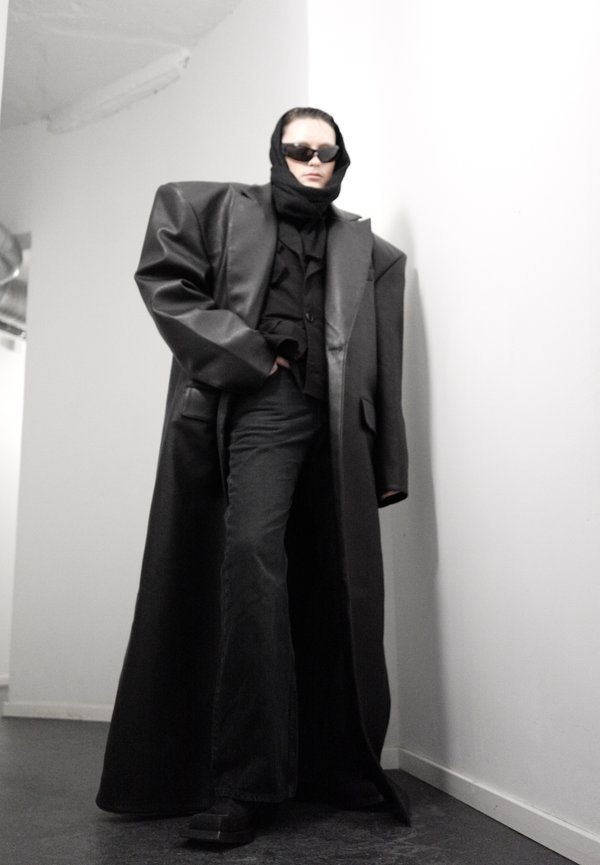 Άτομο που φοράει υπερμεγέθη μαύρο δερμάτινο παλτό, μαύρο μαντήλι στο κεφάλι, γυαλιά ηλίου, μαύρο παντελόνι με φαρδύ πόδι, στέκεται σε μια λευκή γωνία μέσα σε κλειστό χώρο.