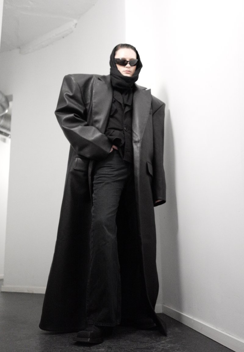 Personne portant un manteau en cuir noir surdimensionné, un foulard noir, des lunettes de soleil, un pantalon noir à jambes larges, debout dans un coin blanc à l'intérieur.