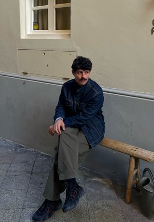 Una persona está sentada en un banco de madera, lleva una chaqueta de mezclilla azul, pantalones verde oliva y zapatos negros con acentos rojos en un ambiente de tonos neutros.