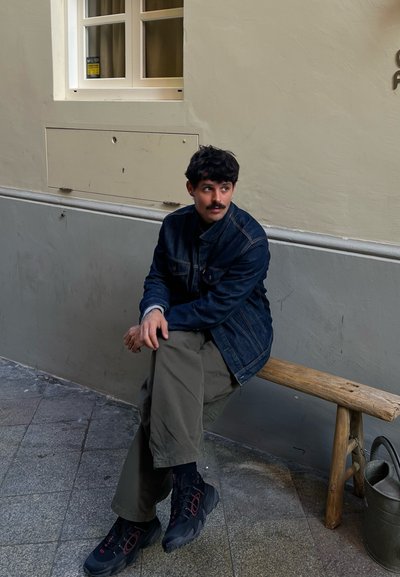 Una persona está sentada en un banco de madera, lleva una chaqueta de mezclilla azul, pantalones verde oliva y zapatos negros con acentos rojos en un ambiente de tonos neutros.