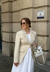 Μπεζ σακάκι προσαρμοσμένο πάνω από ένα λευκό φόρεμα, κρατώντας ένα διάφανο ποτήρι με μια μαύρη καλαμάκι και μια μικρή λευκή τσάντα με χρυσές λεπτομέρειες.