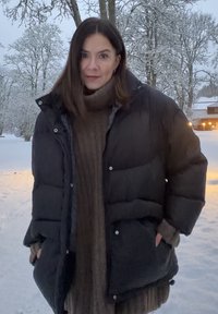 Czarna pikowana kurtka puchowa z teksturą pikowaną i zatrzaskami, nałożona na brązowy sweter z dzianiny, ustawiona na tle zaśnieżonego krajobrazu.