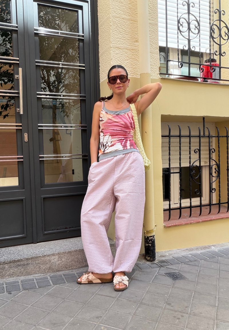 Femme portant des lunettes de soleil, un débardeur coloré, un pantalon large à carreaux roses et des sandales, debout détendue près d'un bâtiment jaune avec une rambarde en métal noir.