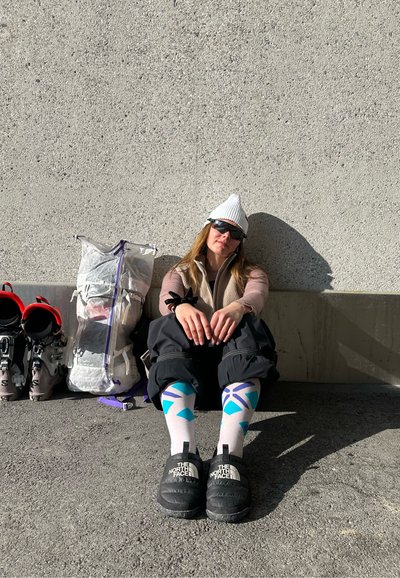 Persona con ropa de invierno y calcetines con patrón sentada contra una pared texturizada, con botas de esquí y una mochila transparente cerca, bajo la luz del sol.