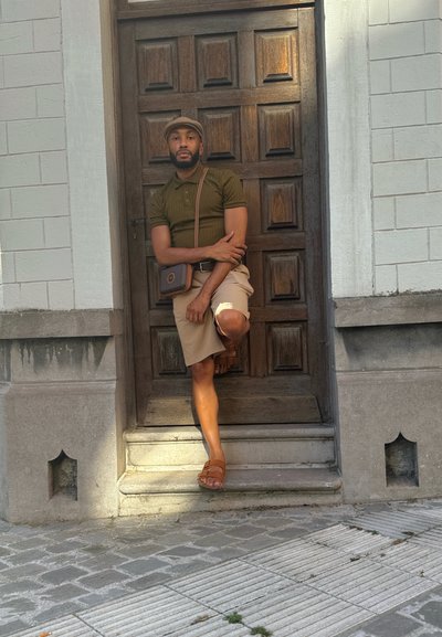Persona apoyada contra una puerta de madera. Lleva una gorra marrón, polo verde oliva, pantalones cortos beige y sandalias marrones. Una bolsa de hombro marrón colgada del hombro.