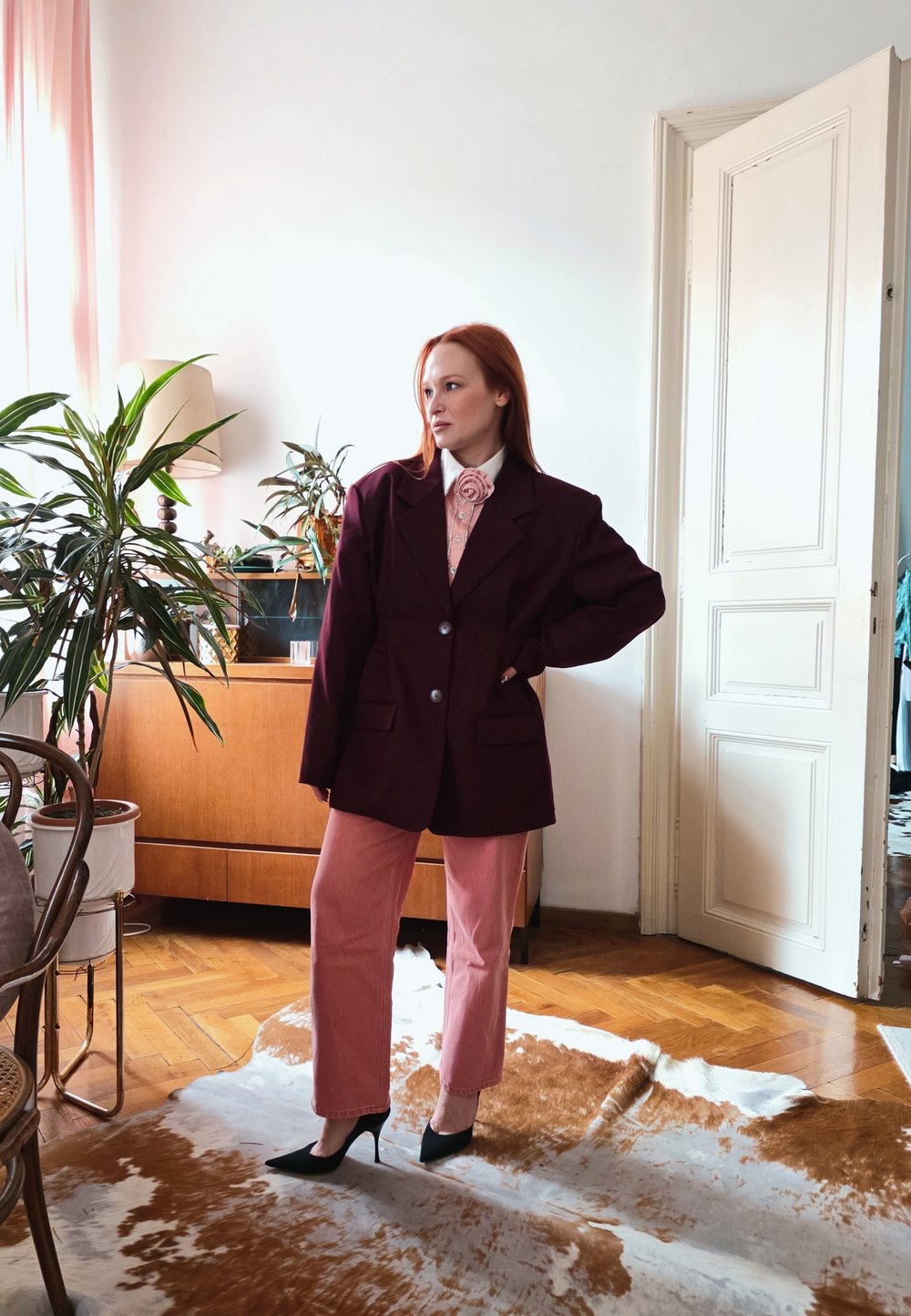 Femme aux cheveux roux portant un blazer noir surdimensionné, un pantalon rose et des talons noirs se tient dans un salon avec des plantes et des meubles en bois.