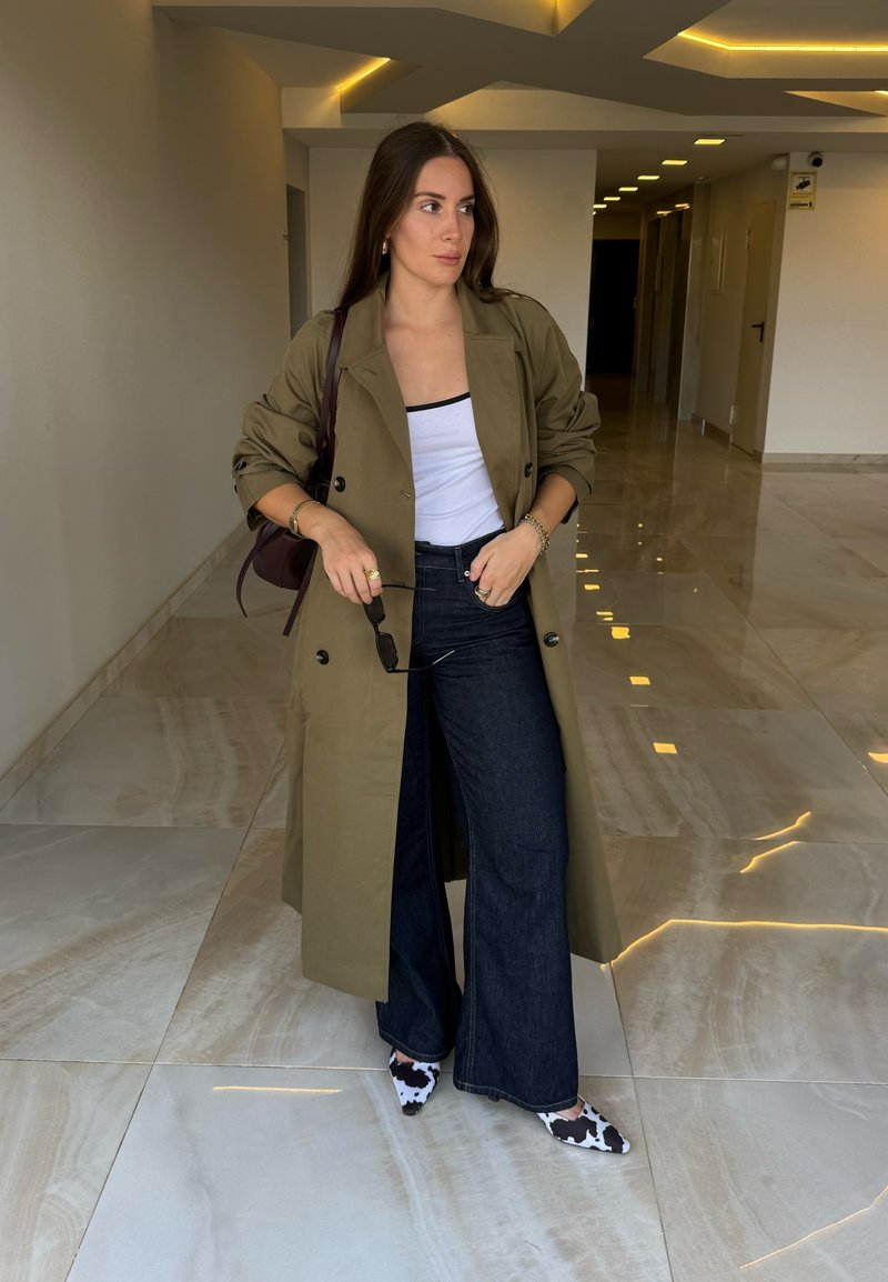 Lungo trench coat verde oliva sopra una maglietta bianca, jeans a zampa d'elefante blu scuro e scarpe con tacco a stampa bovina. In piedi su un pavimento di marmo.