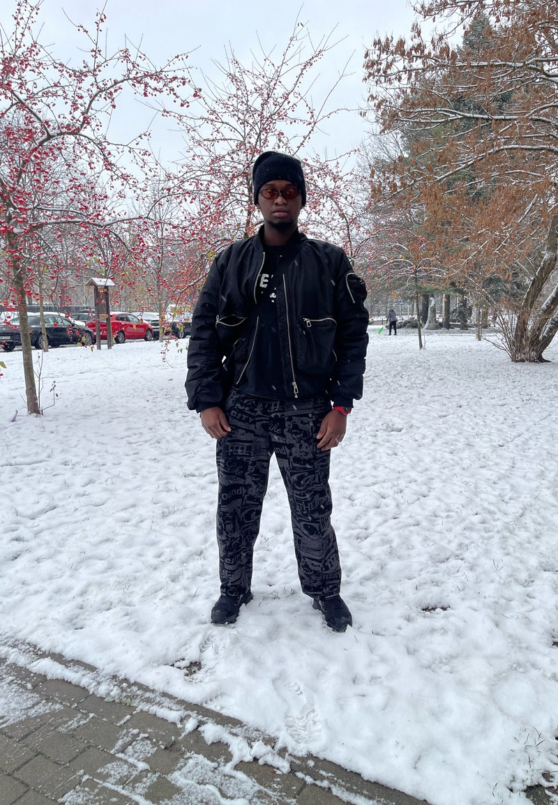 Veste bomber noire avec poches zippées, pantalon graphique noir et chaussures noires. Sol enneigé et arbres avec des baies rouges en arrière-plan.