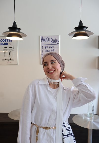 Camisa de lino blanca con mangas de gran tamaño, complementada con un cinturón dorado de diseño y un collar de conchas. El fondo presenta lámparas y decoración.