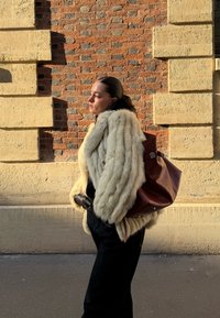 Γυναίκα με μπεζ παλτό από γούνα και μαύρα γάντια στέκεται πλαγίως απέναντι σε έναν τοίχο με υφή από τούβλα και πέτρες, κρατώντας μια μεγάλη καφέ δερμάτινη τσάντα.