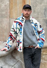 Veste en polaire avec un motif de carte multicolore, assortie à un pull gris et à un jean foncé. La veste possède une fermeture éclair et des poches latérales.