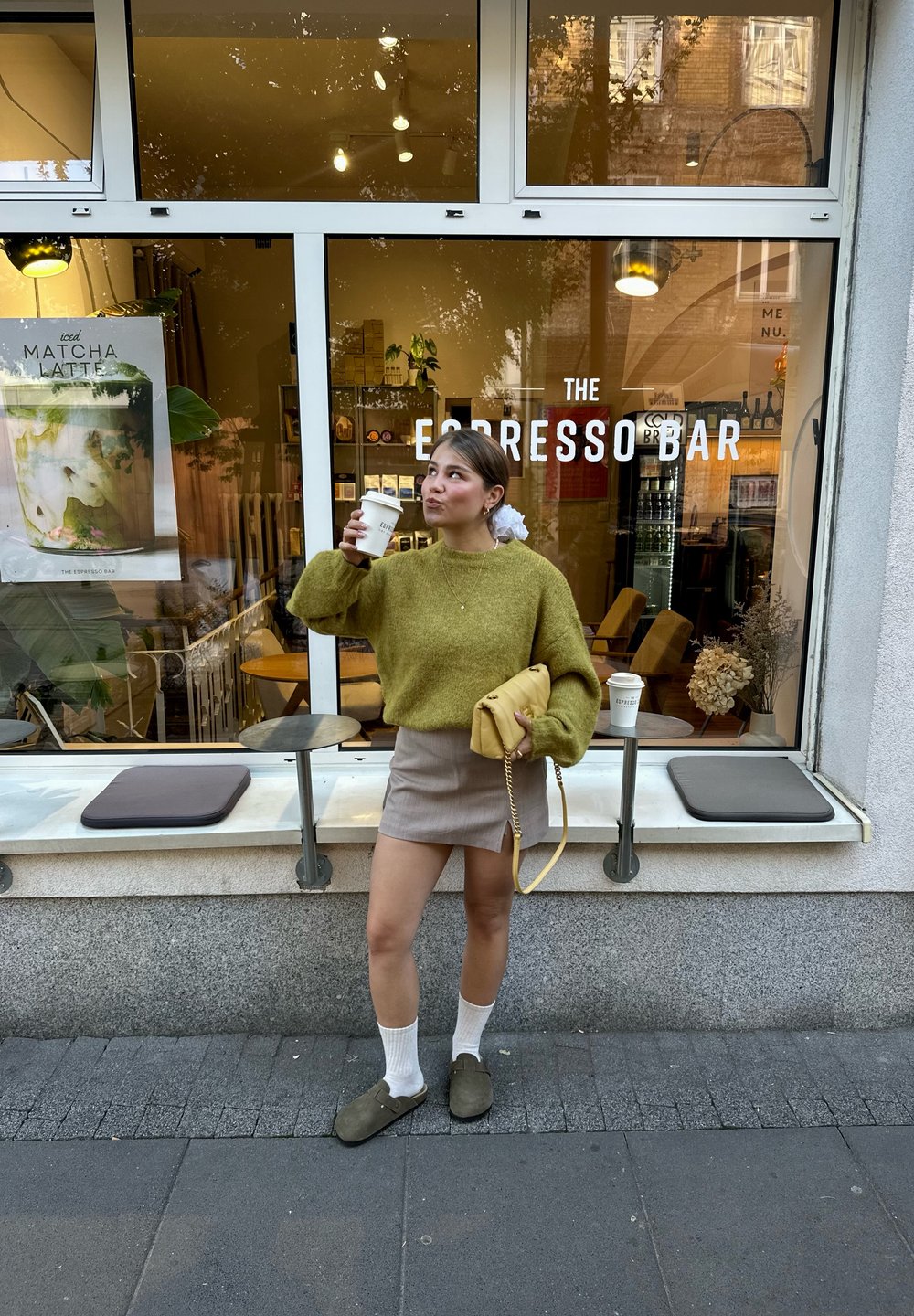 Žena u zelenom džemperu i suknji pije kafu napolju ispred "The Espresso Bar" sa vidljivim mestom za sedenje i posterom menija kroz prozor.