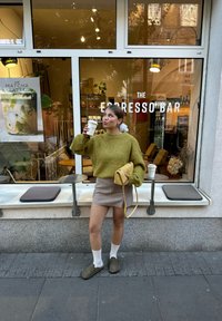 Žena u zelenom džemperu i suknji pije kafu napolju ispred "The Espresso Bar" sa vidljivim mestom za sedenje i posterom menija kroz prozor.