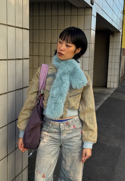 Mujer joven apoyada contra una pared con azulejos, que lleva una bufanda borrosa azul claro, chaqueta beige corta, vaqueros estampados y lleva un bolso morado.