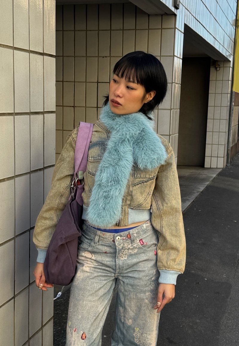 Jeune femme appuyée contre un mur carrelé, portant une écharpe duveteuse bleu clair, une veste beige courte, un jean à motifs et un sac violet.