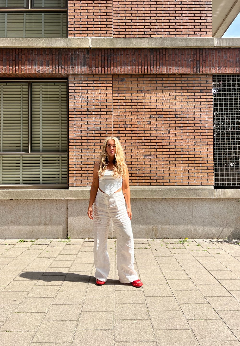 Witte linnen overalls met een getailleerde bodice, brede benen en een subtiele textuur. Gecombineerd met felrode instap schoenen op een betegelde bestrating.