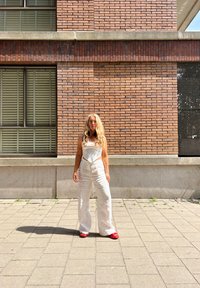 Witte linnen overalls met een getailleerde bodice, brede benen en een subtiele textuur. Gecombineerd met felrode instap schoenen op een betegelde bestrating.