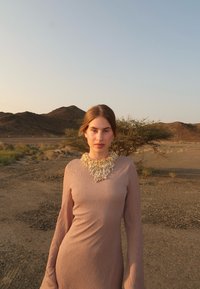 Ung kvinna i en långärmad beige klänning med ett stort, intrikat halsband, stående i ett torrt, bergigt ökenlandskap vid solnedgången.