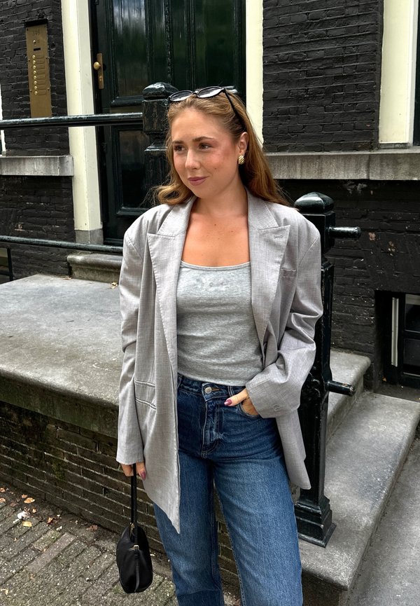 Grijze blazer met gestructureerde schouders over een strak grijze tanktop, gecombineerd met hoge blauwe spijkerbroeken. Aangevuld met een kleine zwarte handtas.