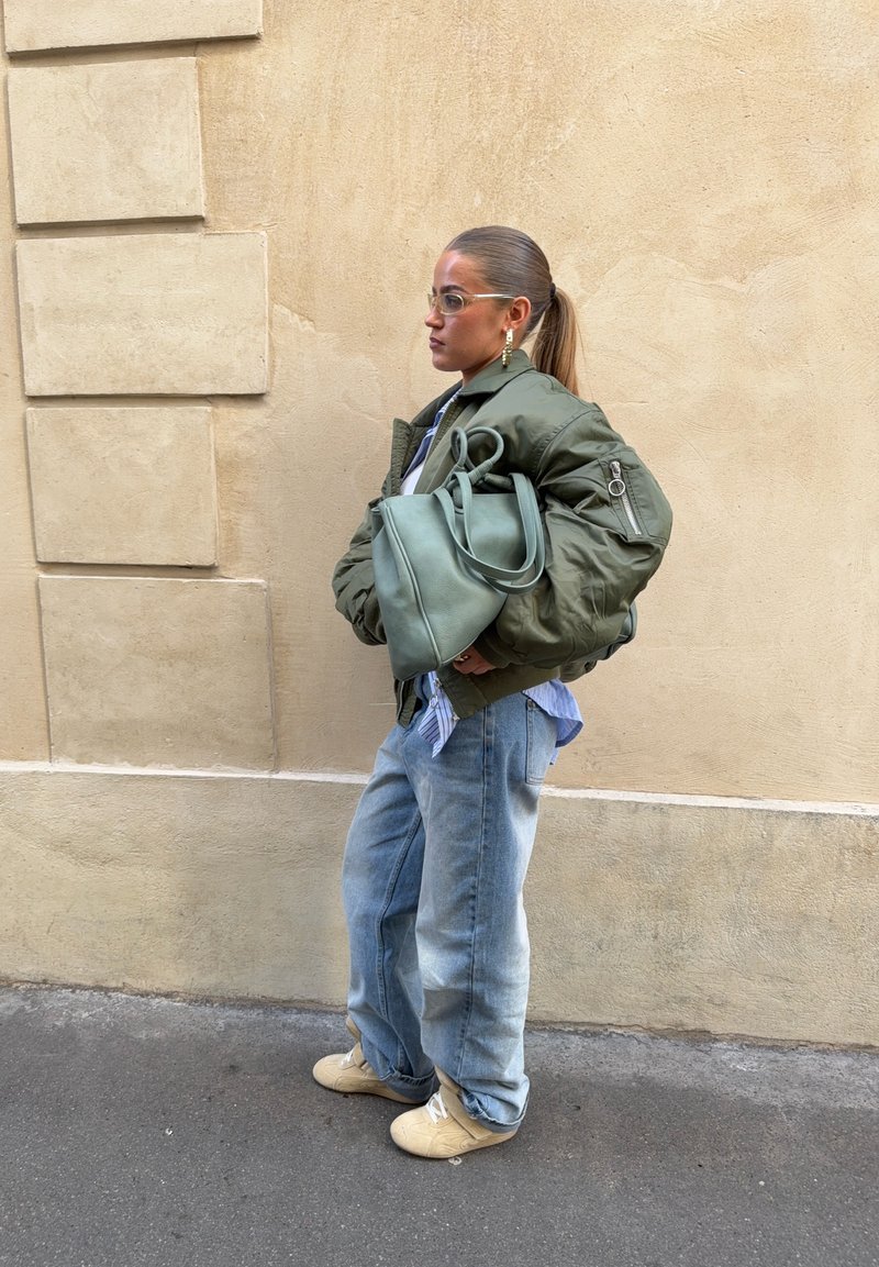 Femme en veste bomber verte surdimensionnée, jean bleu clair, baskets beige, tenant un grand sac à main vert, debout contre un mur beige.