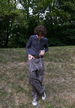 Camisa de manga larga gris, pantalones sueltos de un tono gris marrón con un patrón texturizado y zapatillas verdes. El atuendo incluye un cinturón marrón y tatuajes.